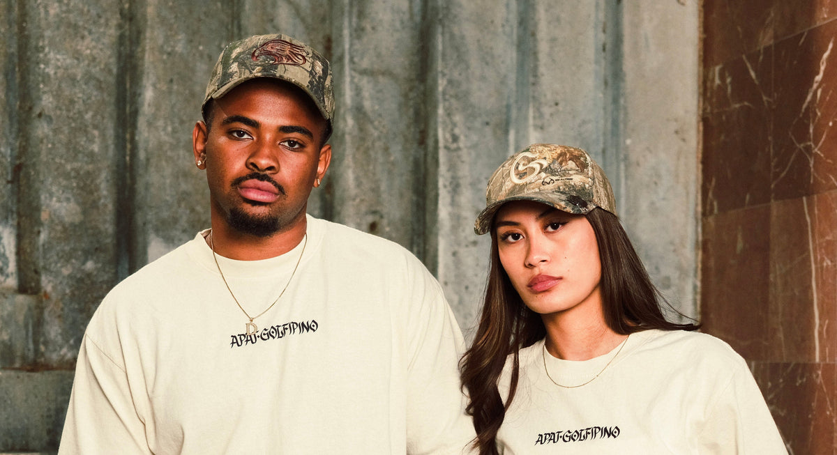 A man and a woman standing side-by-side wearing matching cream-colored t-shirts with black text on the chest and camouflage baseball caps, posed against a textured gray stone wall.