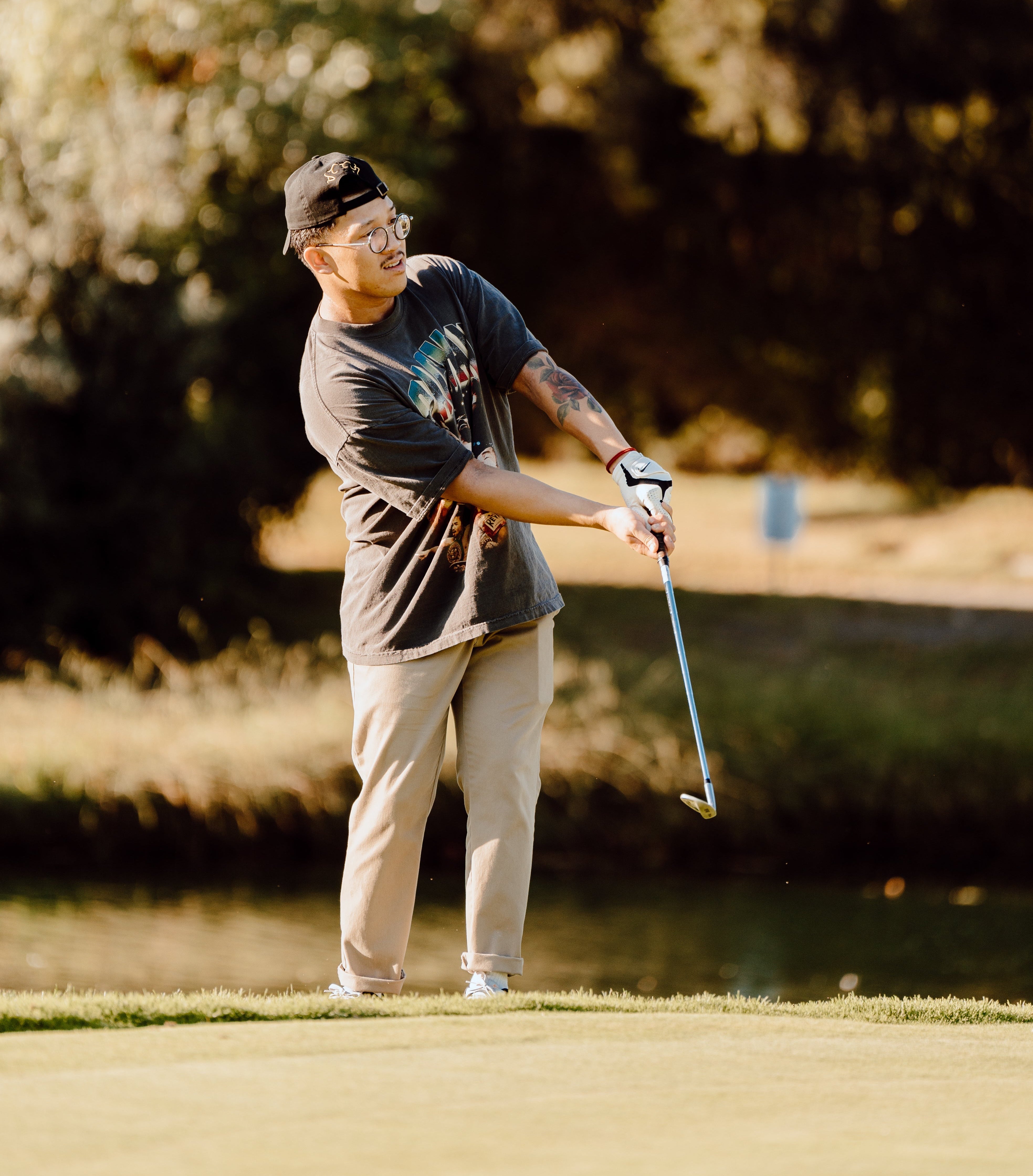 A man wearing a black graphic t-shirt, beige pants, and a black cap is captured mid-swing on a golf course, with trees and water in the background.