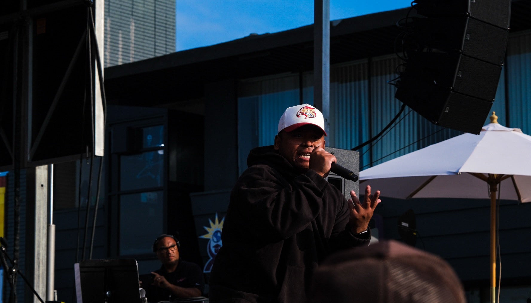 Person speaking into a microphone at an outdoor event with buildings and umbrellas in the background